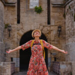 summer trip to Rhodes island, Greece. Young Asian woman in ethnic red dress walks over one of gates Rhodes fortress, road over moat. female traveler visiting southern Europe Unesco world heritage site